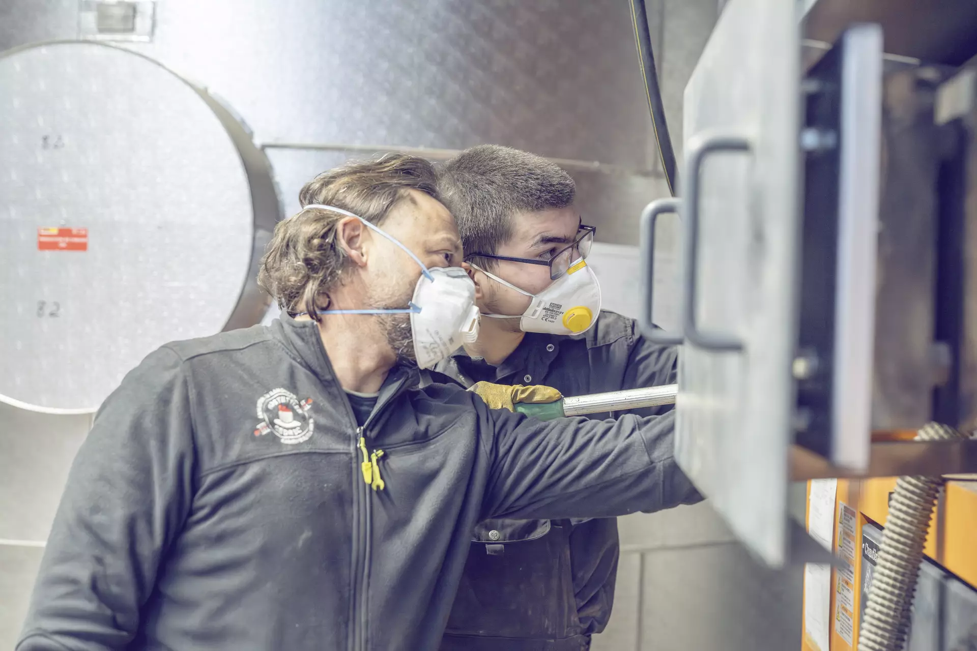 Apprenti ramoneur et son formateur en train de regarder à l'intérieur d'une cheminée industrielle. Les deux ont un masque sur la bouche et des lunettes de protections