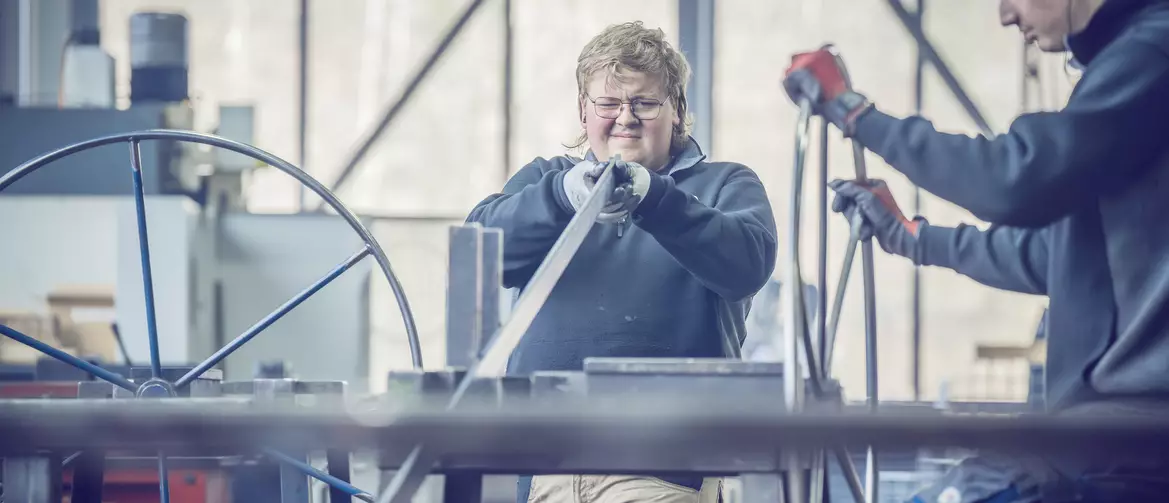 constructeur métallique fermant un oeil pour examiner une longue pièce de métal. Sur la droite une autre personne tient un cercle de métal ressemblant à un gouvernail