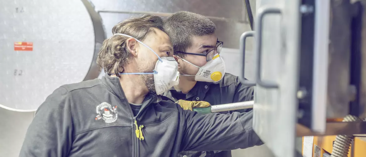 Apprenti ramoneur et son formateur en train de regarder à l'intérieur d'une cheminée industrielle. Les deux ont un masque sur la bouche et des lunettes de protections